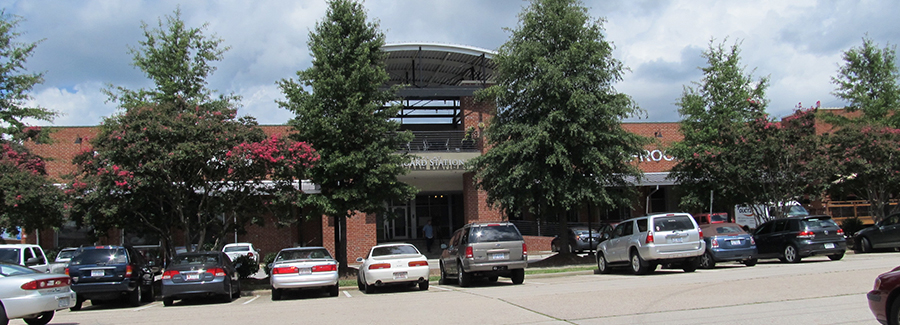Two-story brick building with an upper balcony, mature trees, and a parking lot filled with cars, green and brick-red tones